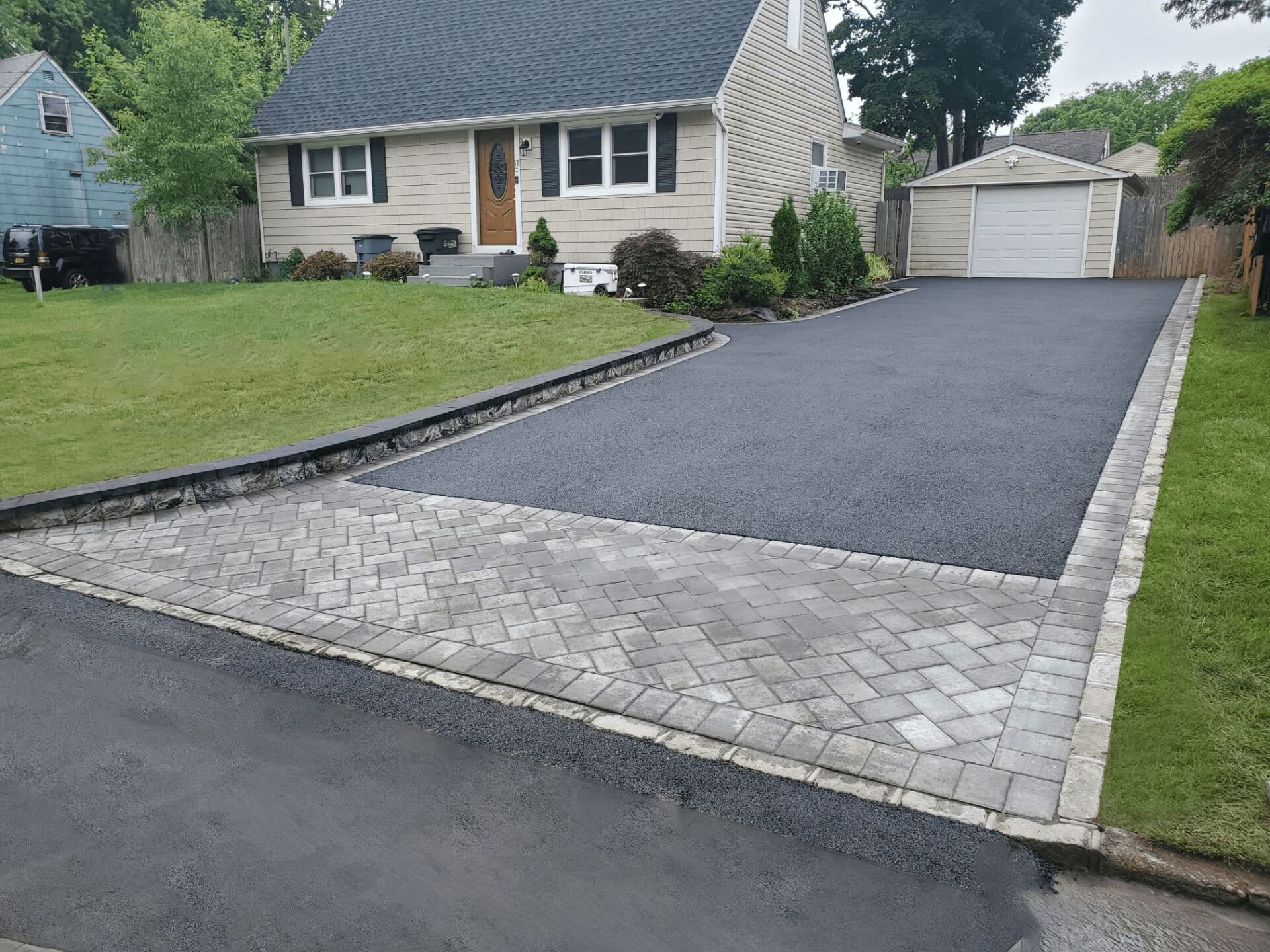 Asphalt driveway with brick pavers leading to a beige house with green lawn.