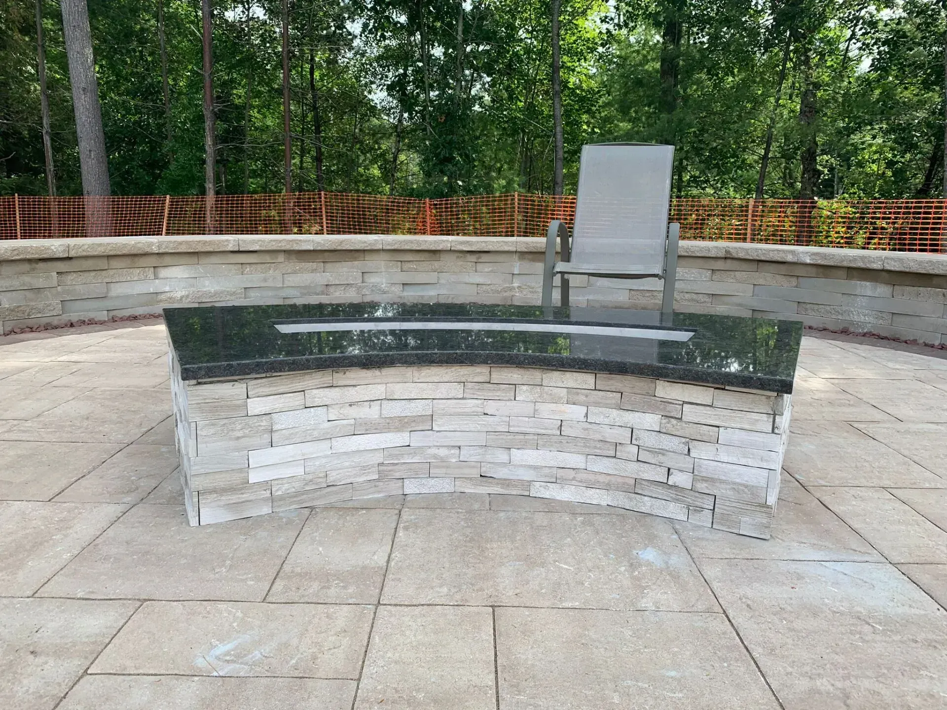 Curved stone bench with a black top and a light gray chair on a patio with a forest backdrop.