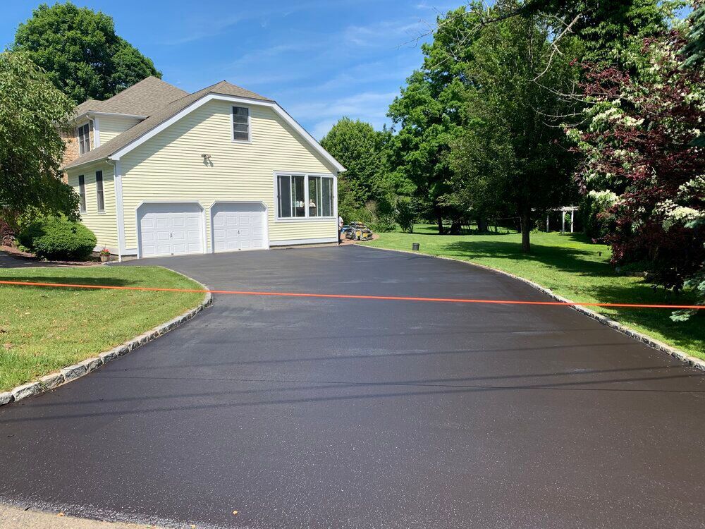 Freshly paved black asphalt driveway in front of a yellow house with a green lawn on a sunny day.