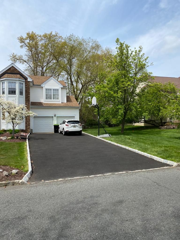 White house with paved driveway, car parked, basketball hoop, trees, and blue sky.