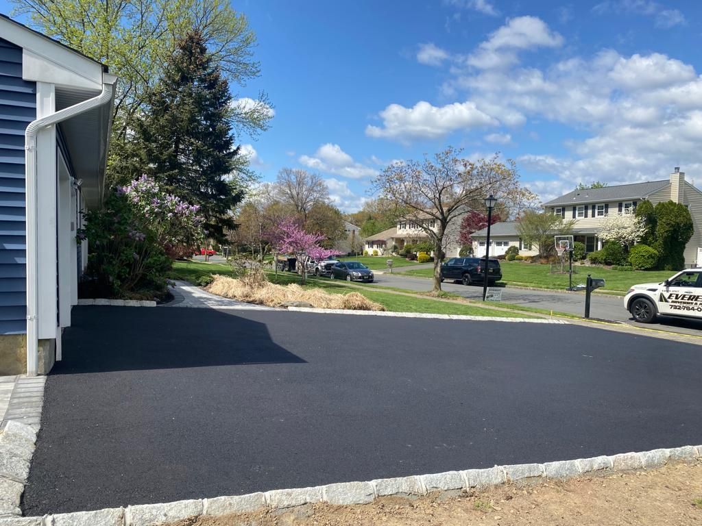 Newly paved black asphalt driveway bordered with stone. Houses and trees in background on a sunny day.