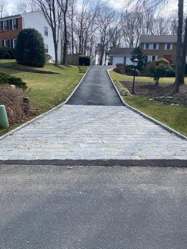 Driveway leading uphill, with a paved section transitioning to a gray brick section. Green grass borders the sides.