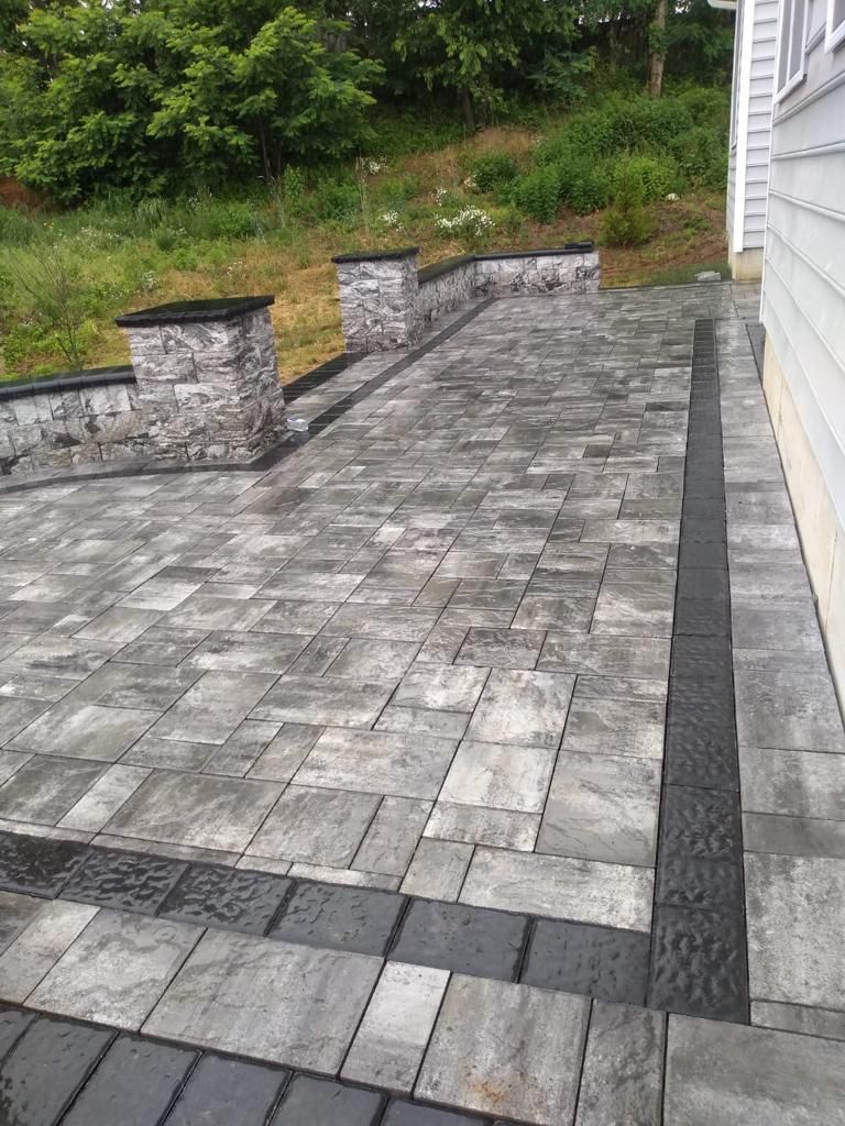 Gray paver patio with stone accents and two pillars, next to a light-colored house.