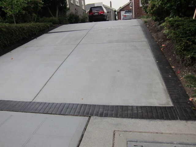 Concrete driveway leading uphill with a dark brick border and cars parked at the top.