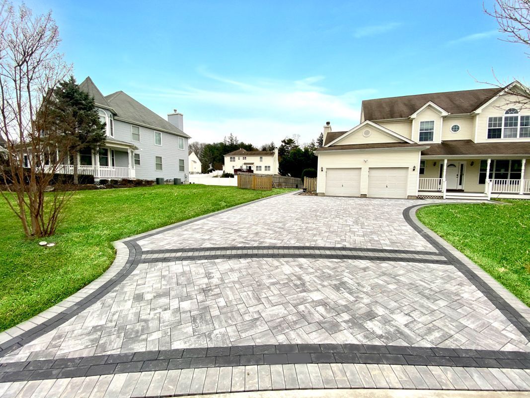 Driveway paved with gray and black bricks leading to a two-story house on a sunny day.