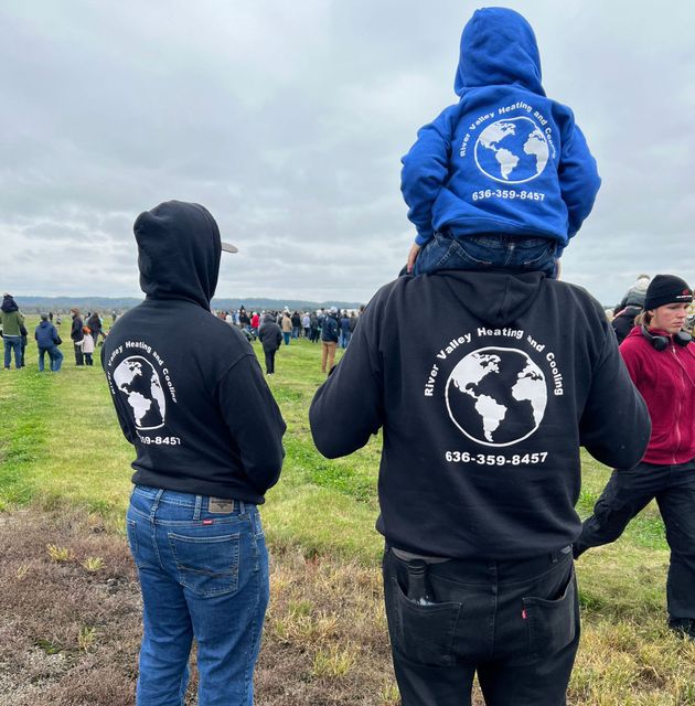 People in hoodies stand in a grassy field; one holds a child on their shoulders.