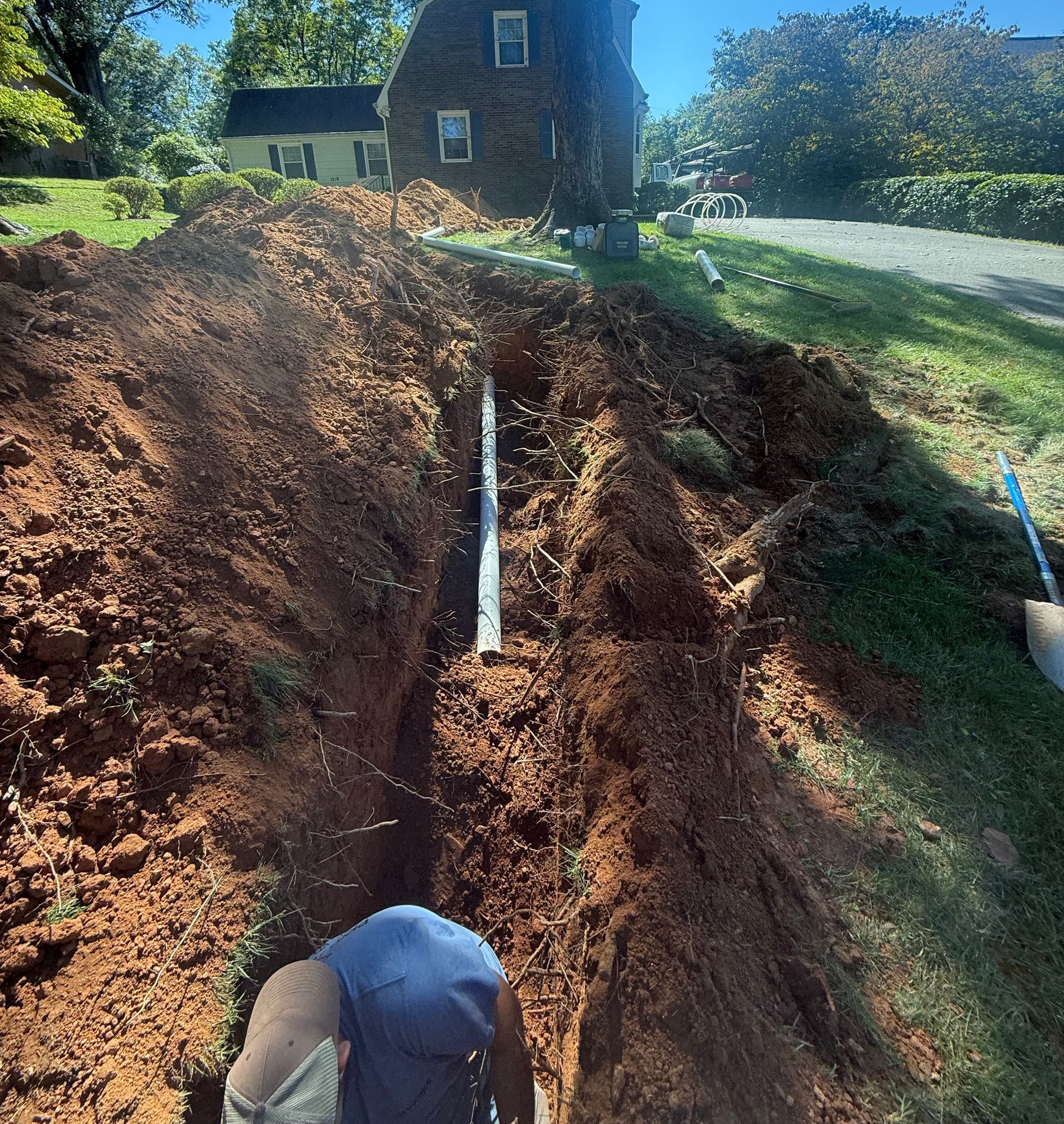 A person in a ditch installing pipe, near a house and grassy area.