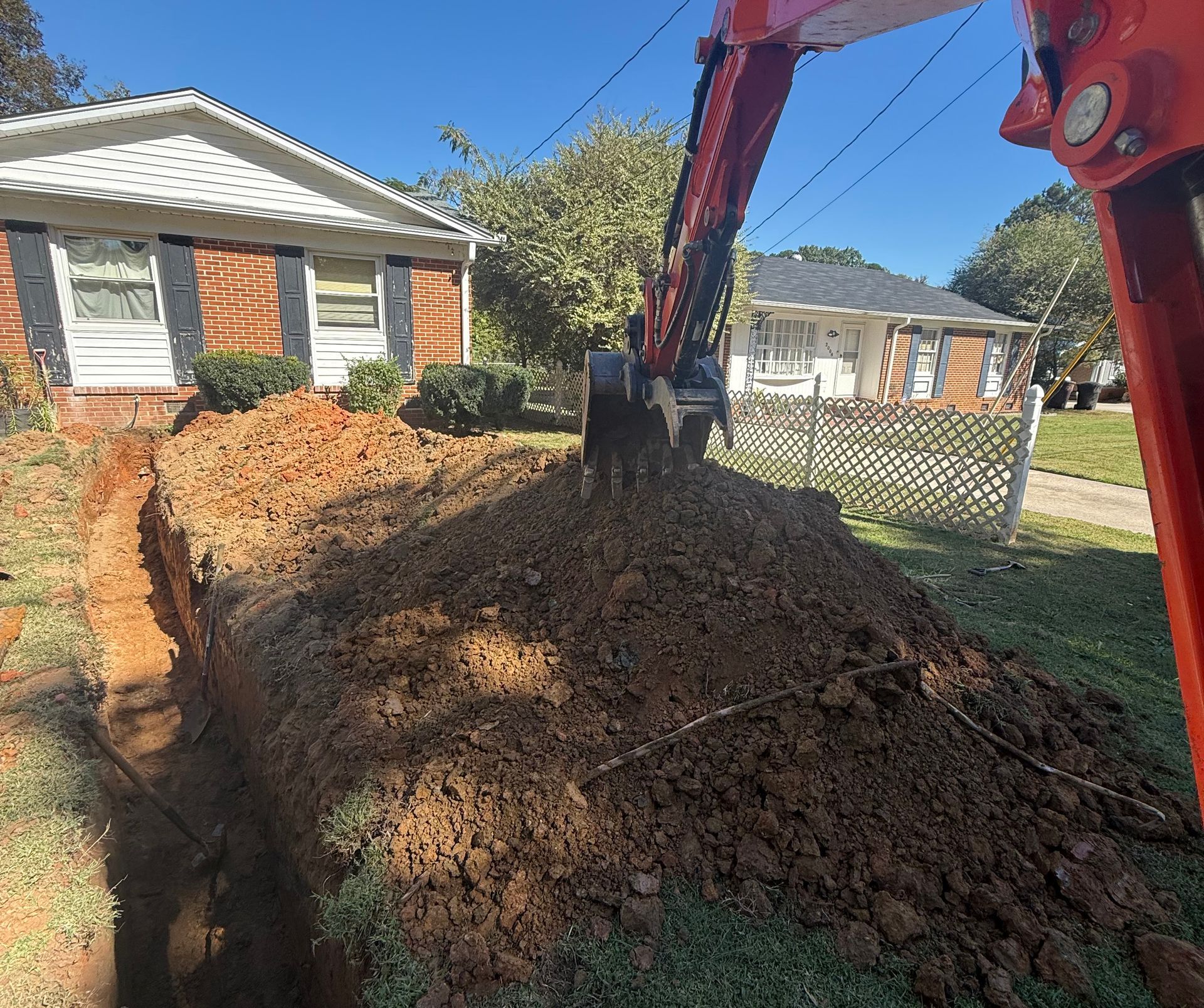 An excavator digging a trench in front of a brick house, creating a large pile of dirt.