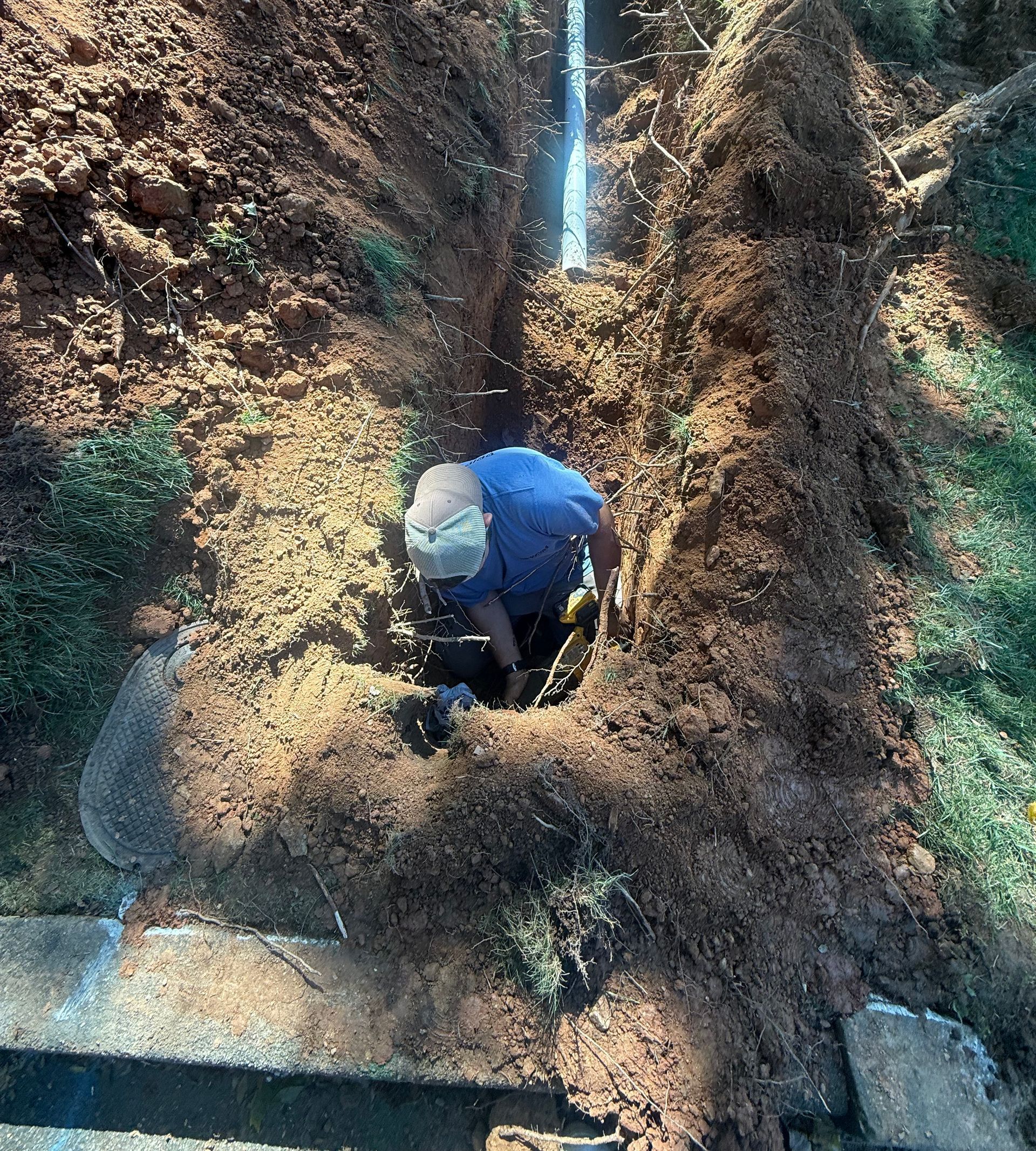 Person in blue shirt working in a deep trench, possibly repairing a pipe. Earth walls and a pipe visible.