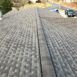 A high-angle view of a grey shingled roof with a dark ridge cap, looking down towards a driveway with a dark truck.