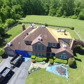 An aerial view of a house undergoing a roof replacement, with workers on the roof, bundles of shingles, and a trailer below.