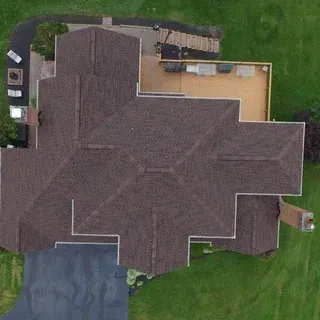 An aerial view of a brown-shingled residential house roof, a paved driveway, a wooden deck, and a surrounding grass lawn.