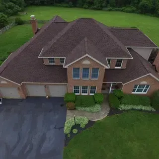 An aerial view of a two-story brick house with a dark shingled roof, a paved driveway, and surrounding green lawn.
