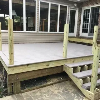 A partially finished wooden deck with light gray planks, four vertical support posts, and steps leading to a patio.