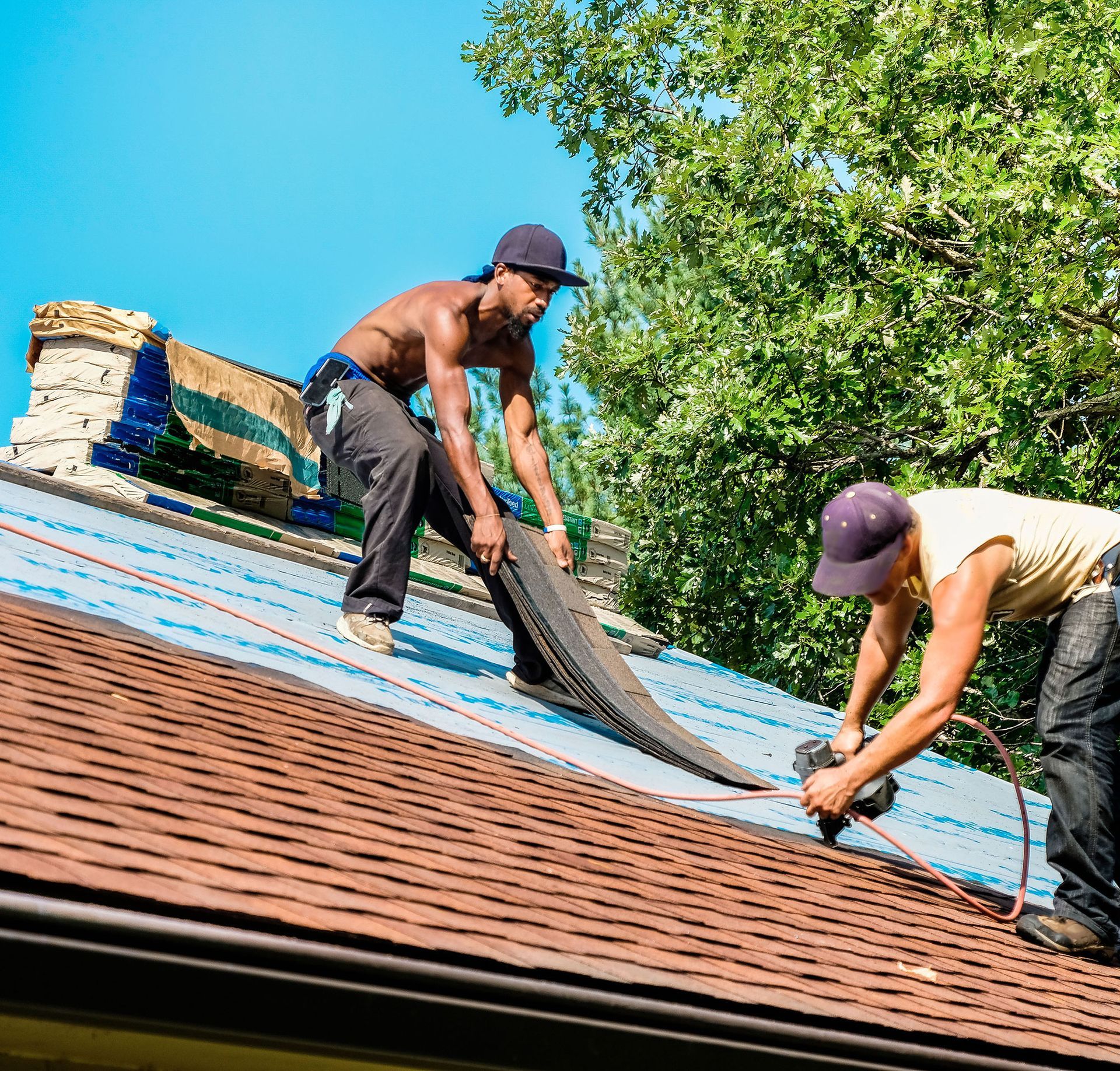Two workers installing blue roofing underlayment on a sloped roof near trees and a bright blue sky