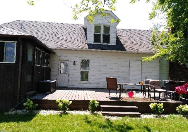 A back view of a white, two-story house with a wooden deck, outdoor seating, and a dormer window on a sunny day.
