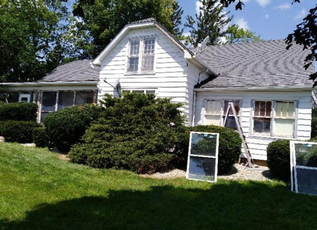 A white, two-story house with a grey roof, surrounded by bushes, with a ladder and removed windows resting against it.