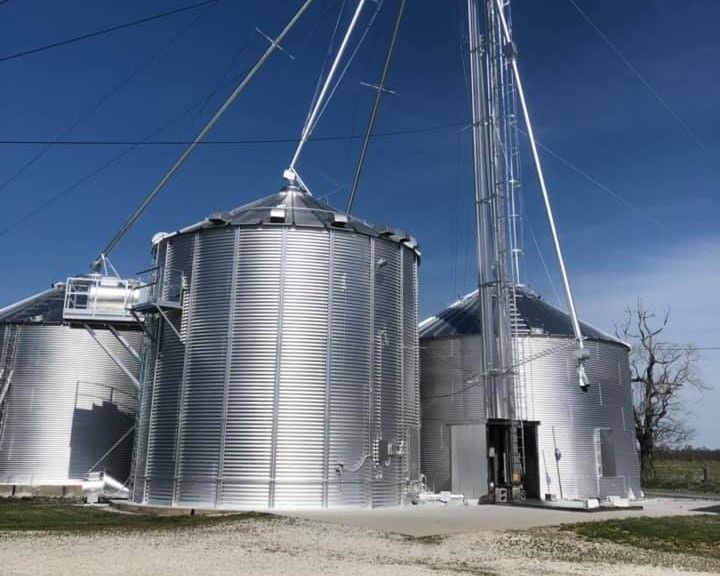 Three large, shiny metal grain silos on a sunny day.  A tall elevator tower is next to one.