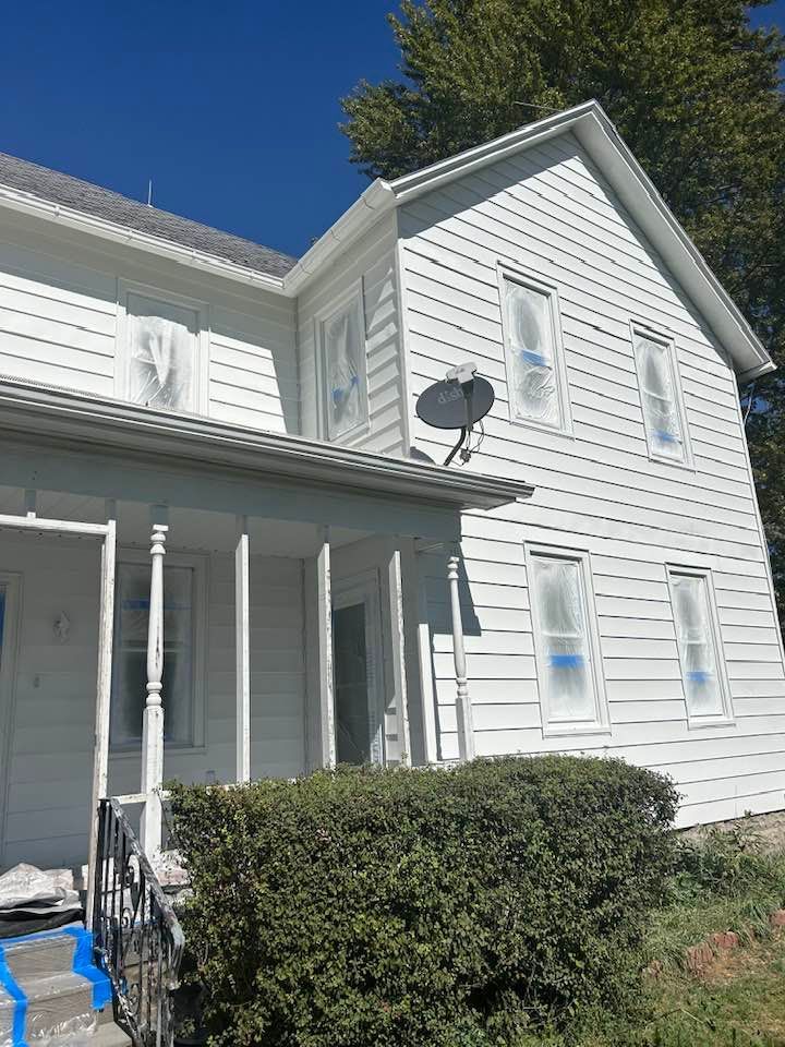 White house being painted, windows taped off, with a porch and shrubbery.