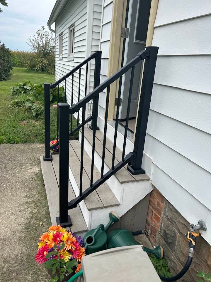 Black handrail and steps leading to a white house entrance. Flowers and watering cans are at the base.