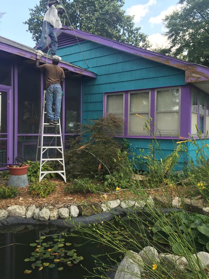 Two people on a ladder and roof, working on a teal and purple house next to a pond.