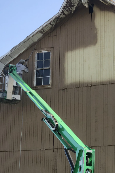 Man on a green lift painting the side of a brown building with a window.
