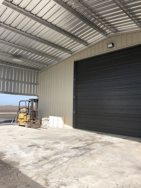 A gray metal-roofed warehouse interior with a closed black door, concrete floor, and a forklift.