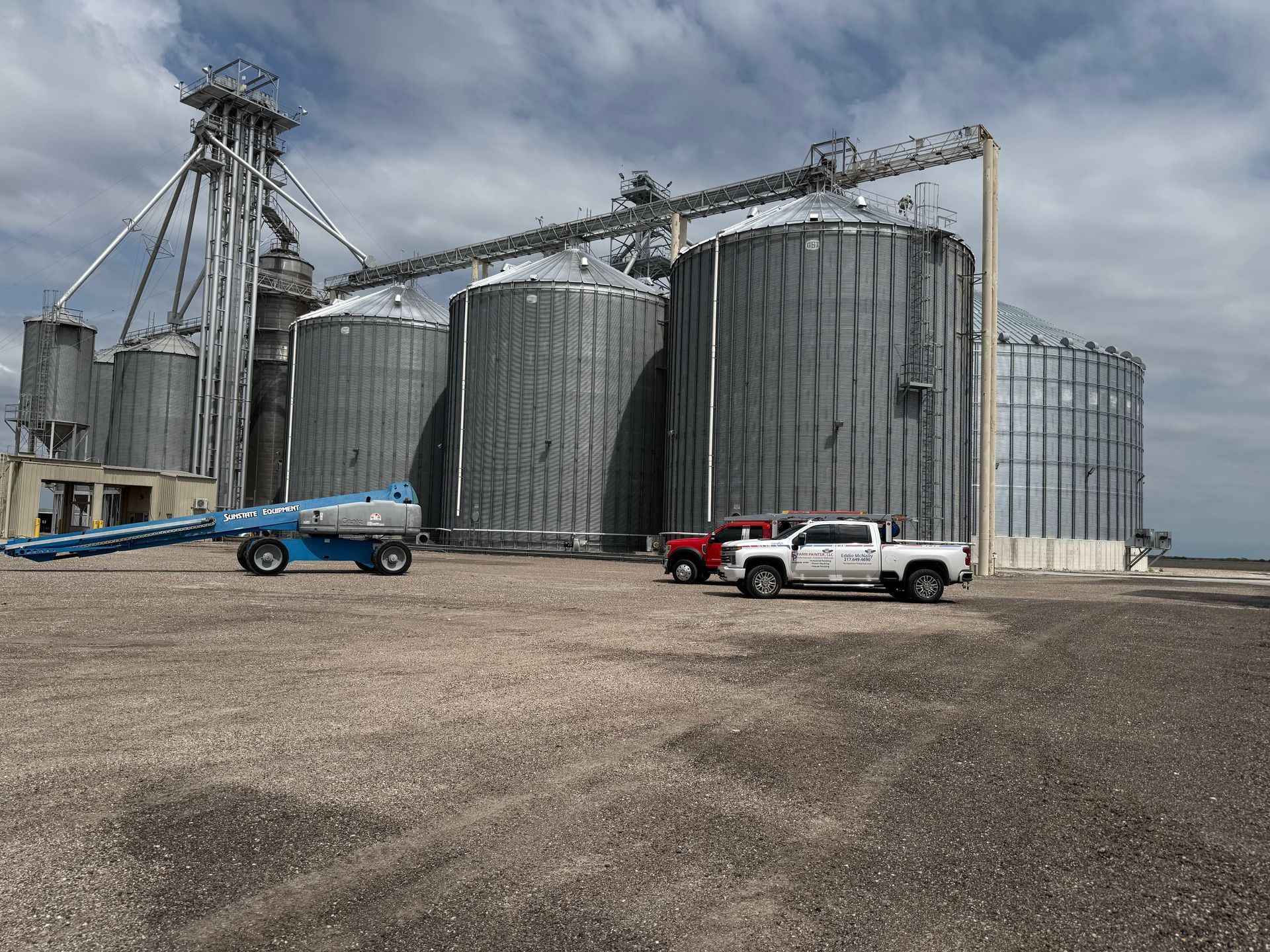 Grain silos, truck, and lift sit on a gravel lot under a cloudy sky.