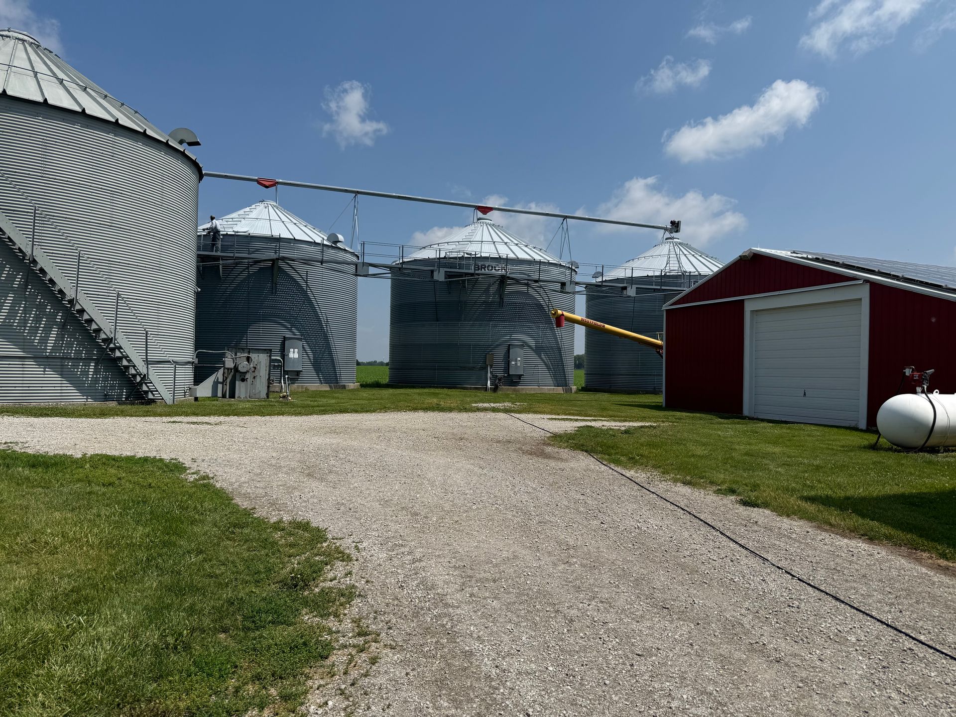 Grain silos and a small red building on a farm, gravel driveway and blue sky.
