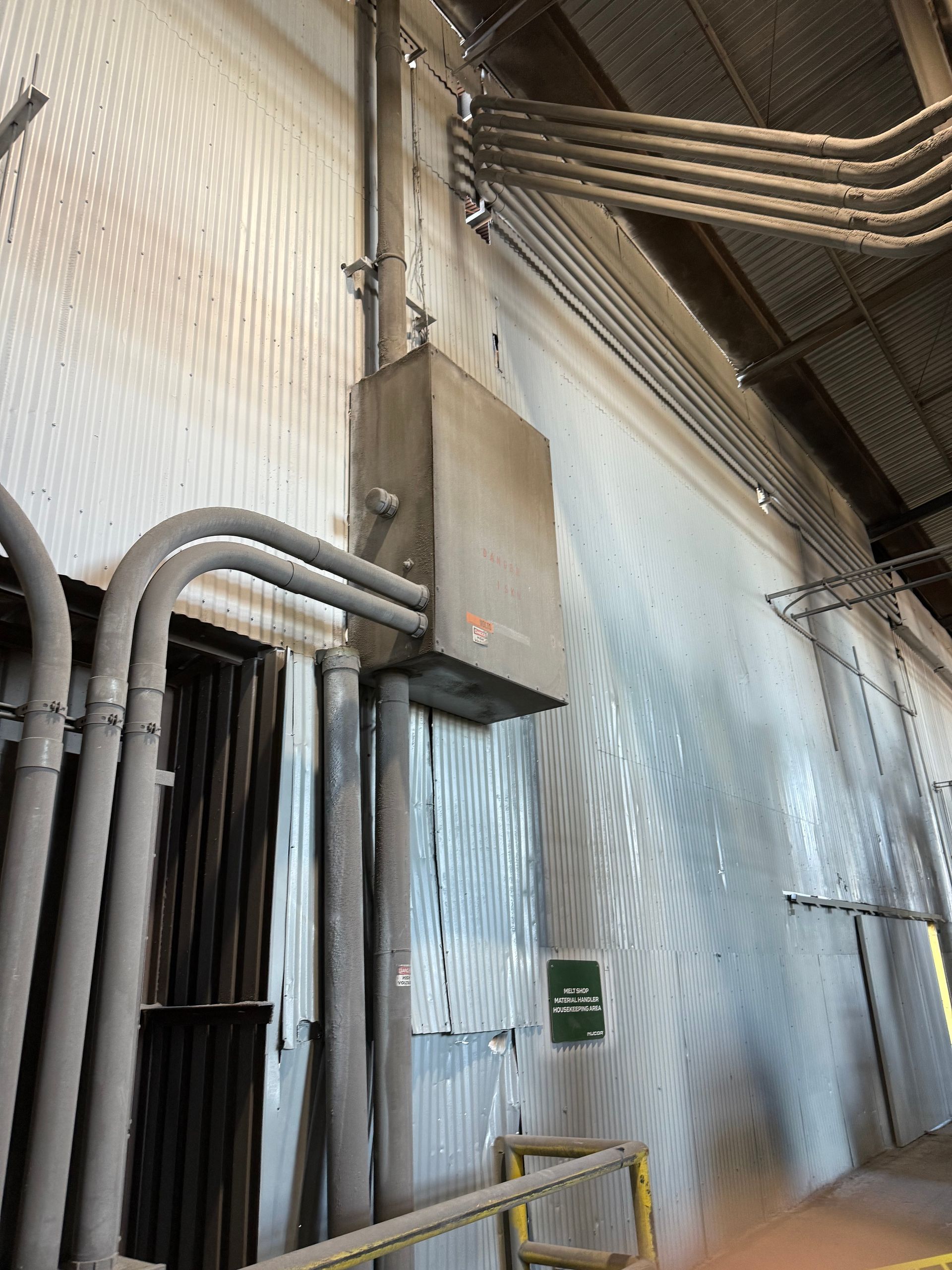 Electrical box and conduit mounted on a corrugated metal wall. Grey pipes and wires are visible.