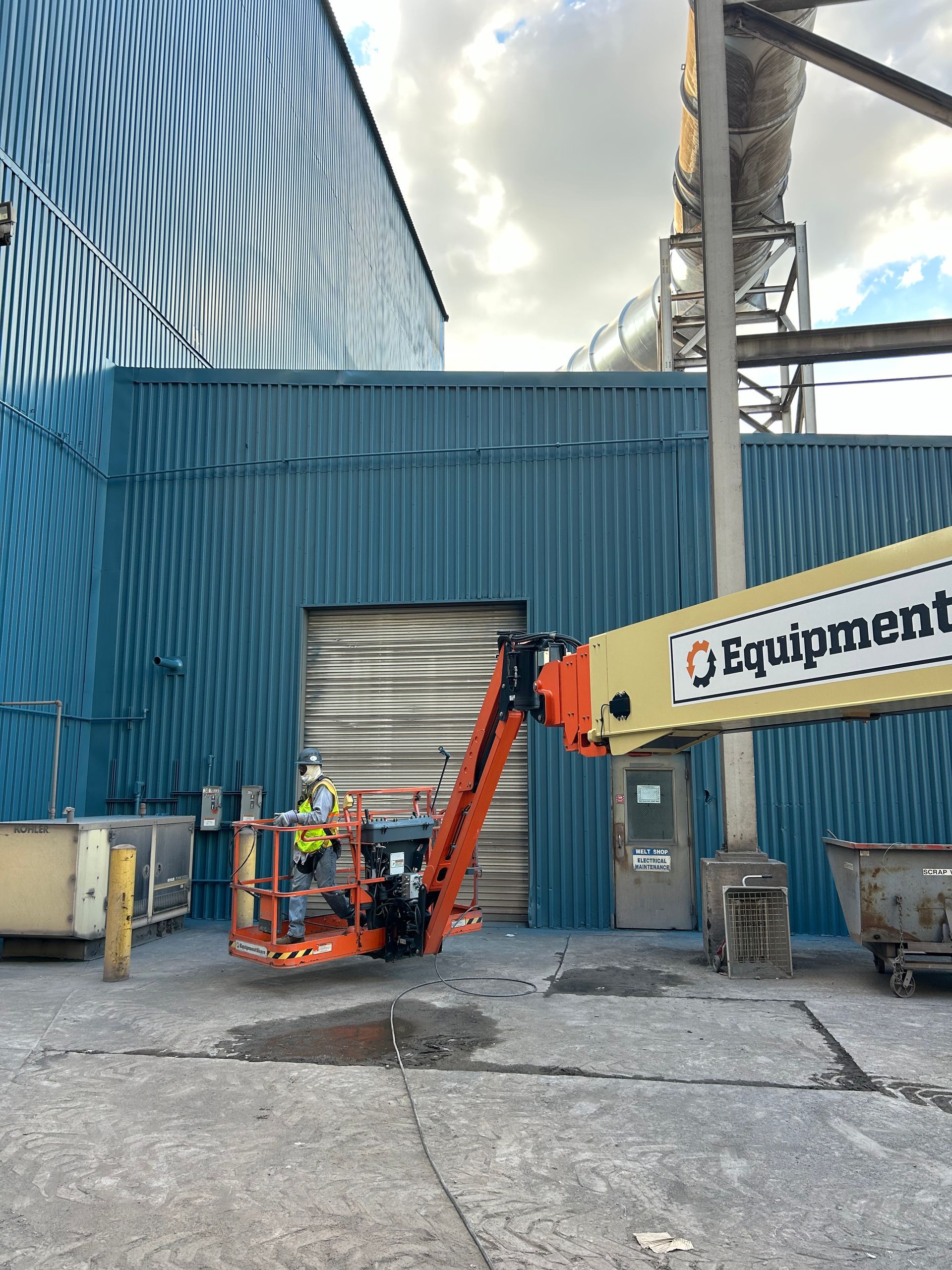 Worker in an orange lift near a blue industrial building, working on the side.
