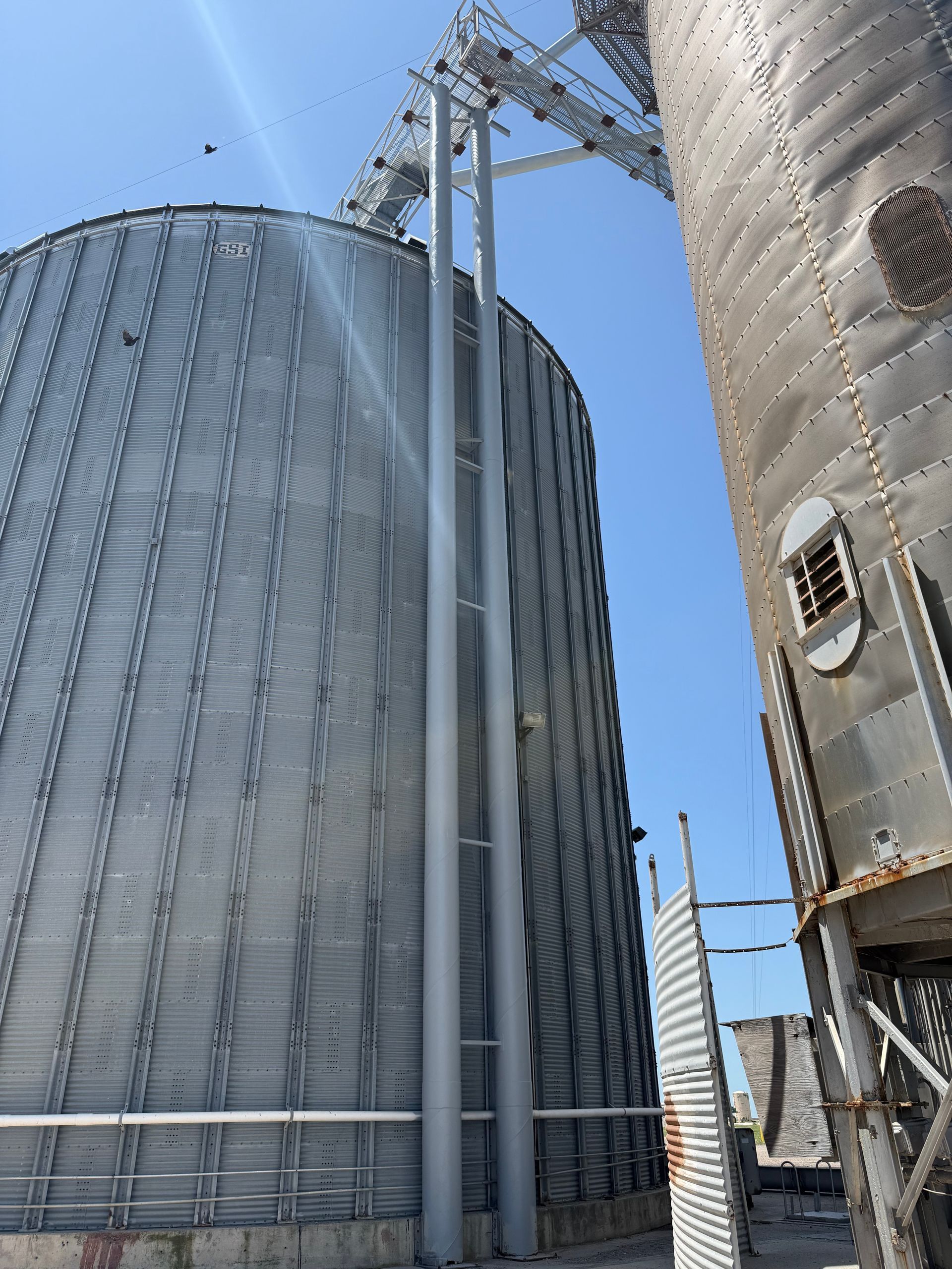 A large metal grain silo stands under a clear blue sky, connected to a smaller structure by an overhead conveyor system.