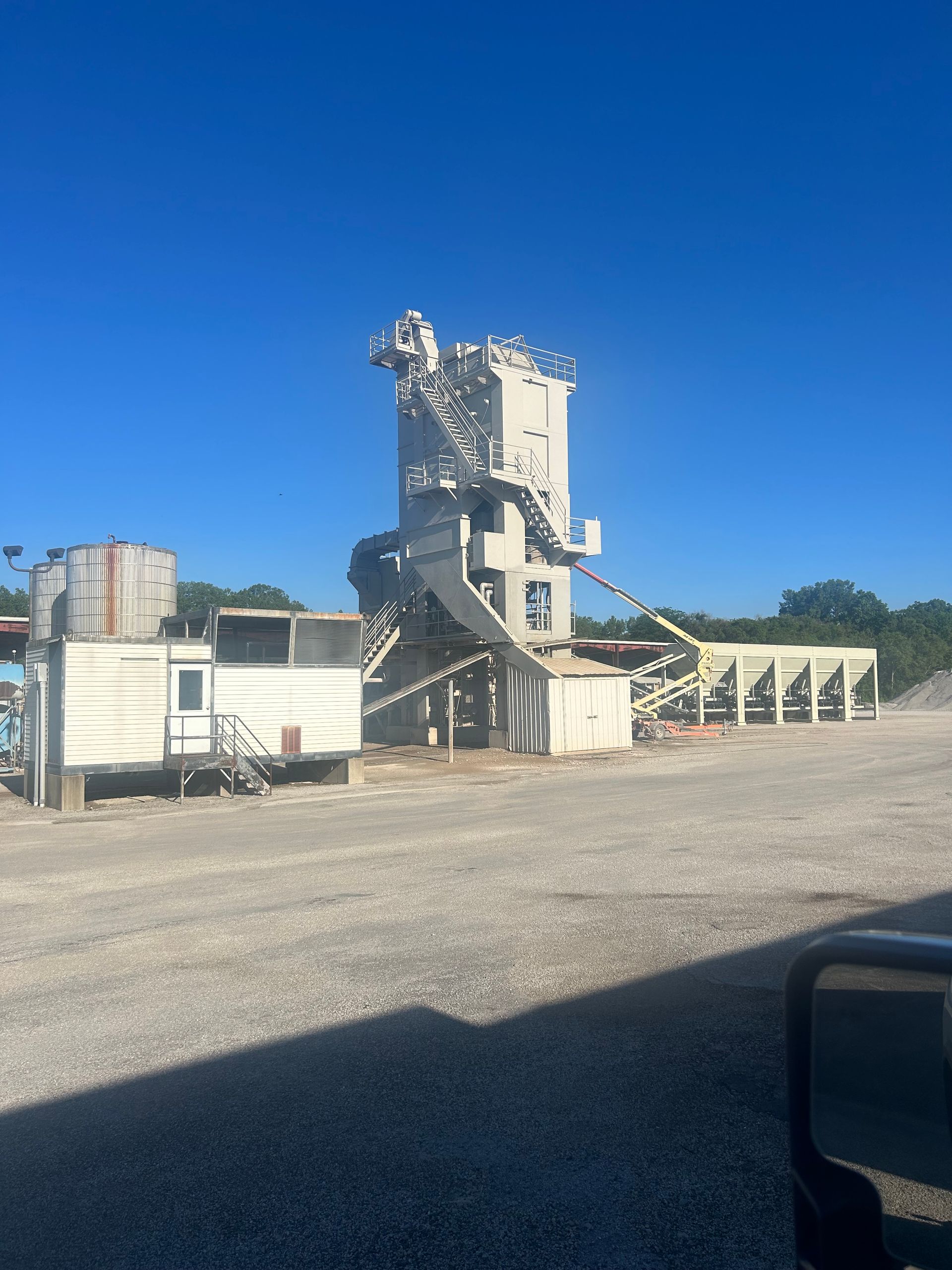 A concrete batch plant with a central tower, conveyor belt, and adjacent office building under a clear blue sky.