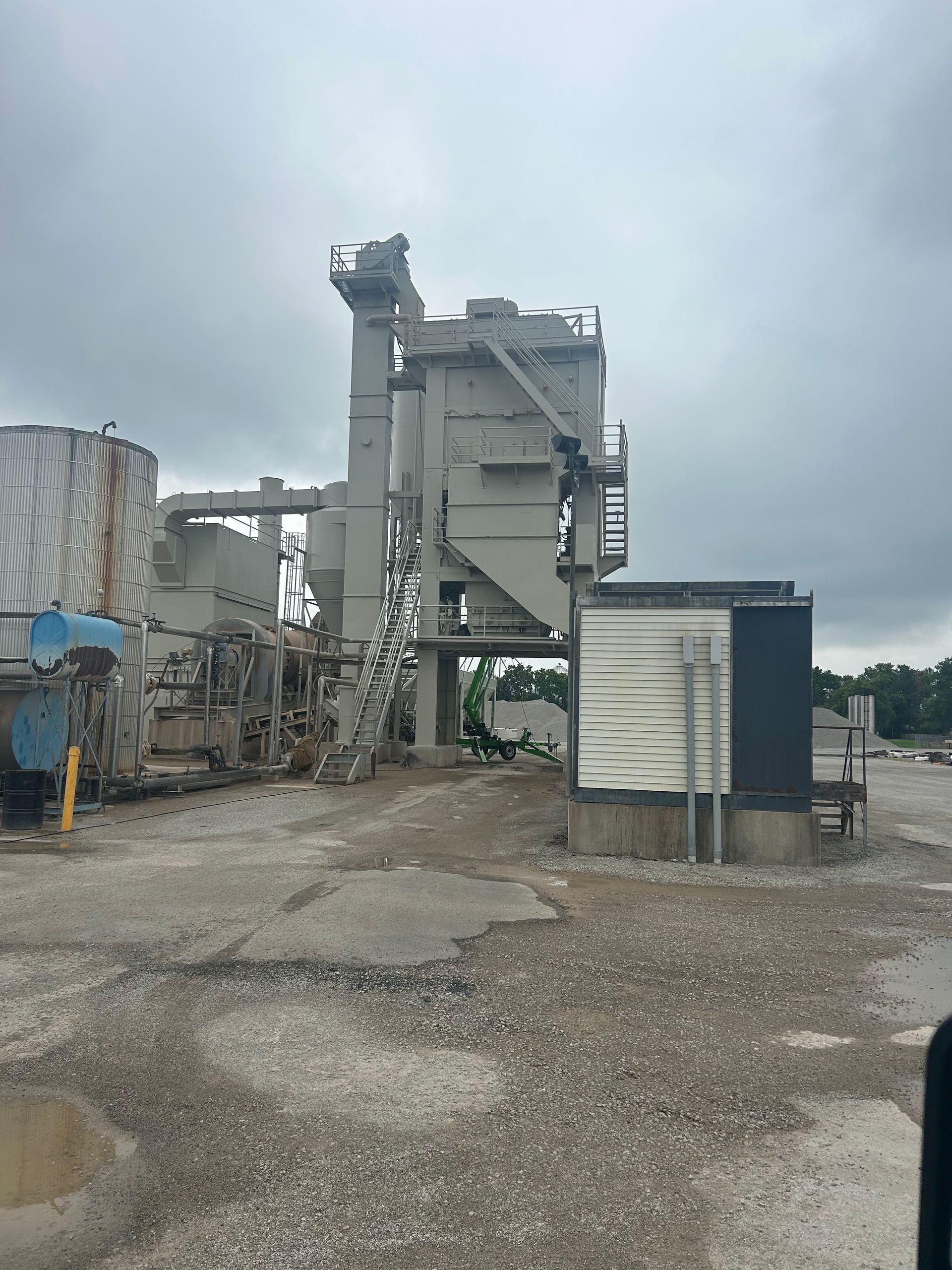 A tall, industrial facility with metal structures and silo-like towers against a cloudy, gray sky over a gravel ground.