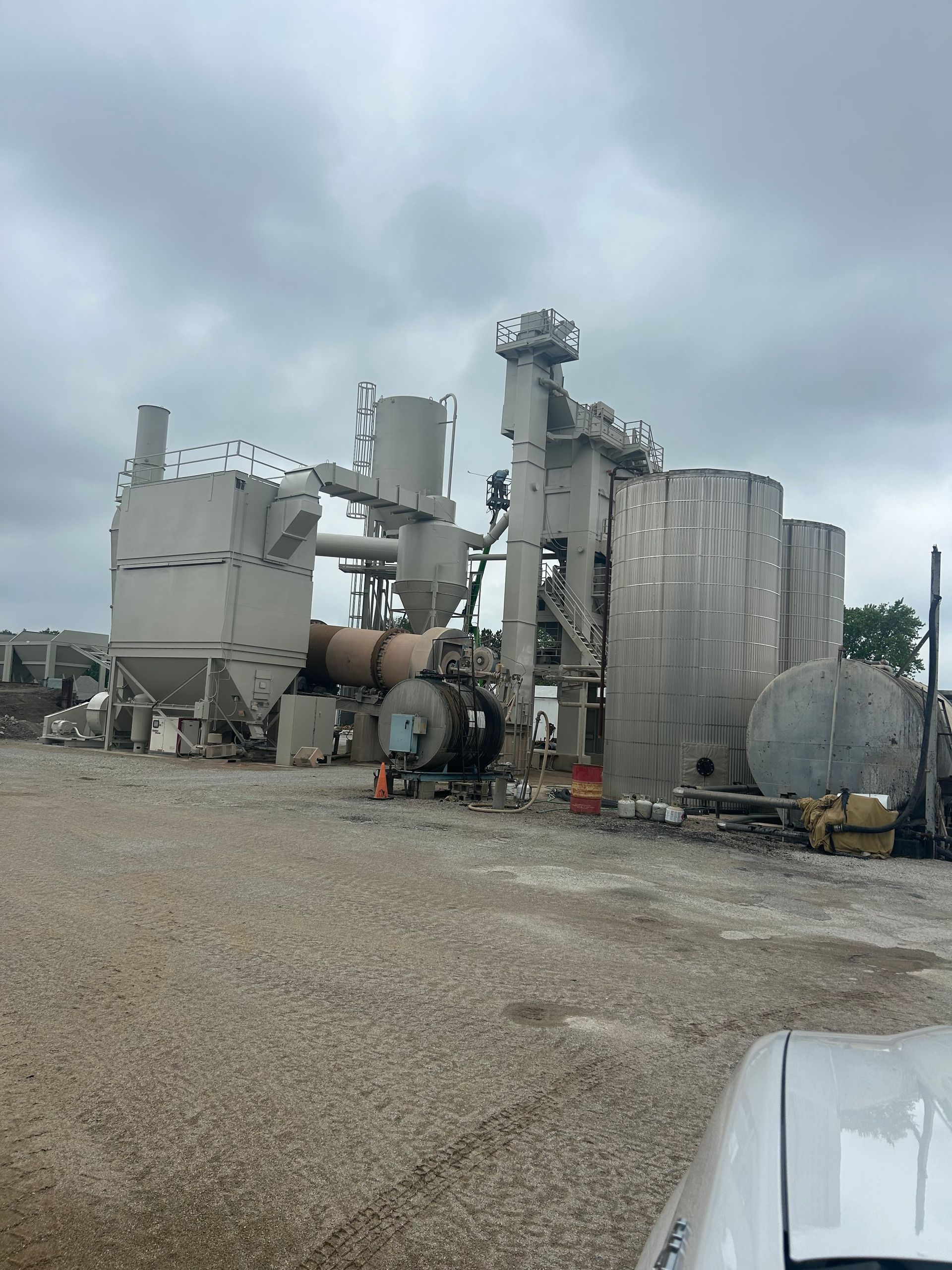 A tall, industrial asphalt processing plant structure made of grey metal sits on a gravel lot under a cloudy sky.
