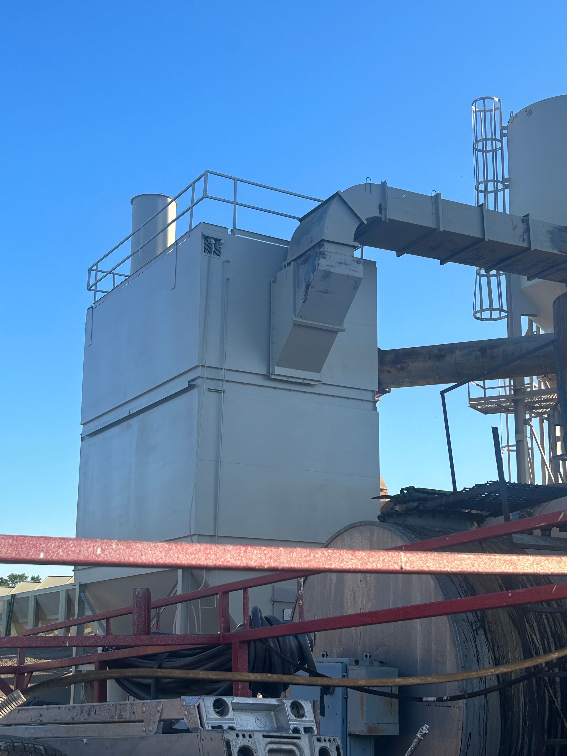 A gray metal industrial dust collector sits outdoors against a clear blue sky, connected by a large overhead duct.