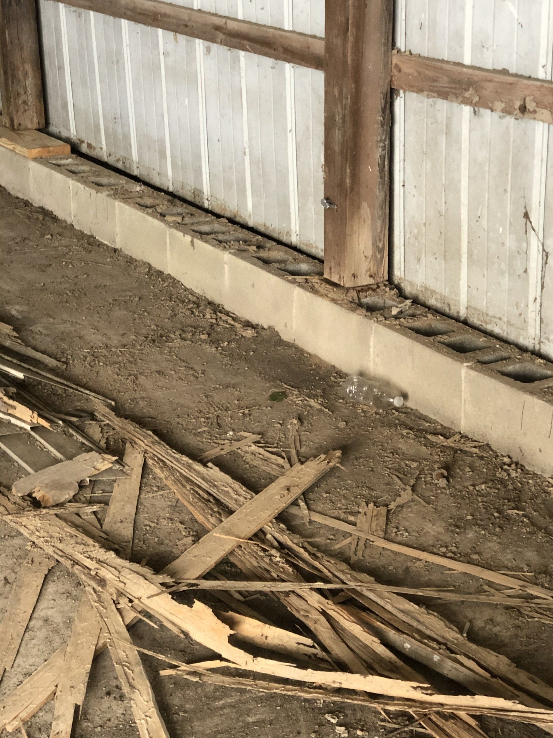 A wooden post and metal siding sit atop a concrete block wall above a floor covered with piles of wood debris.