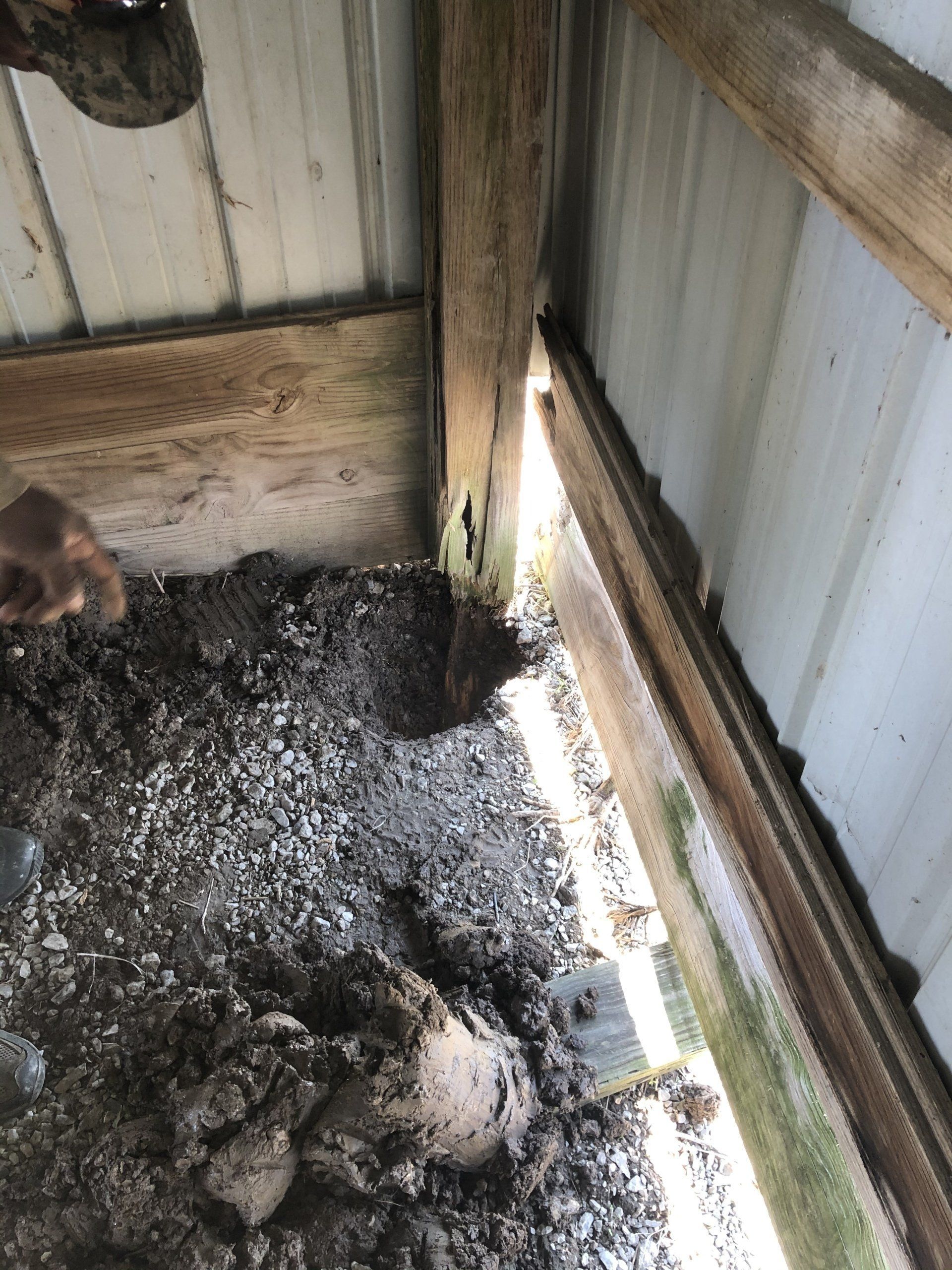 A corner of a barn with a hole in the mud floor, a rotting wooden post, and light shining through an exterior wall gap.