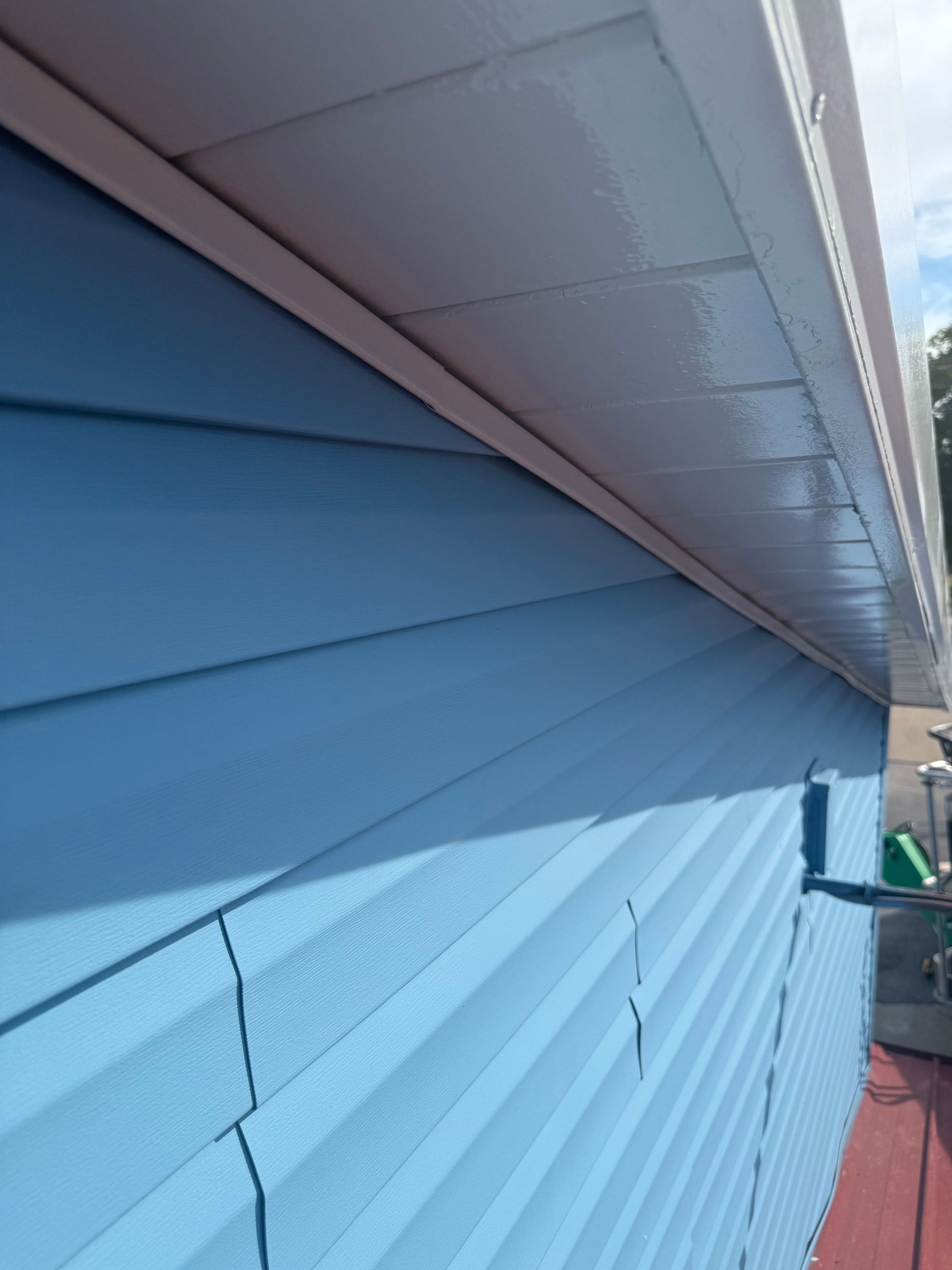 A blue-sided house with white trim being painted by a roller on an extension pole.