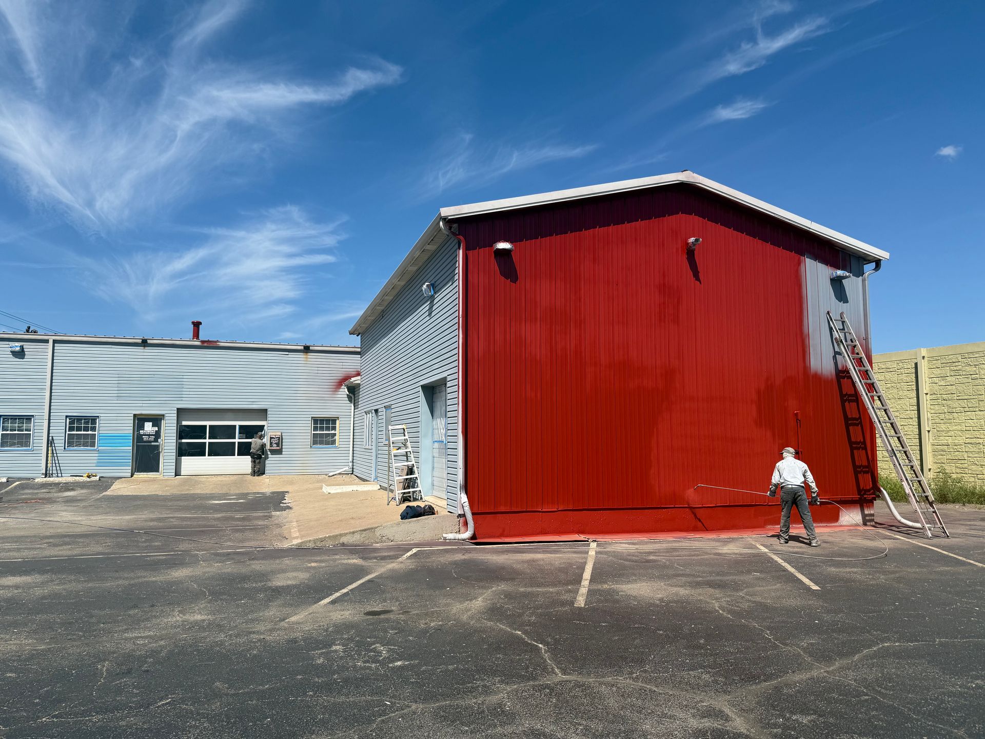 A person uses a ladder to paint a building wall bright red under a sunny blue sky.