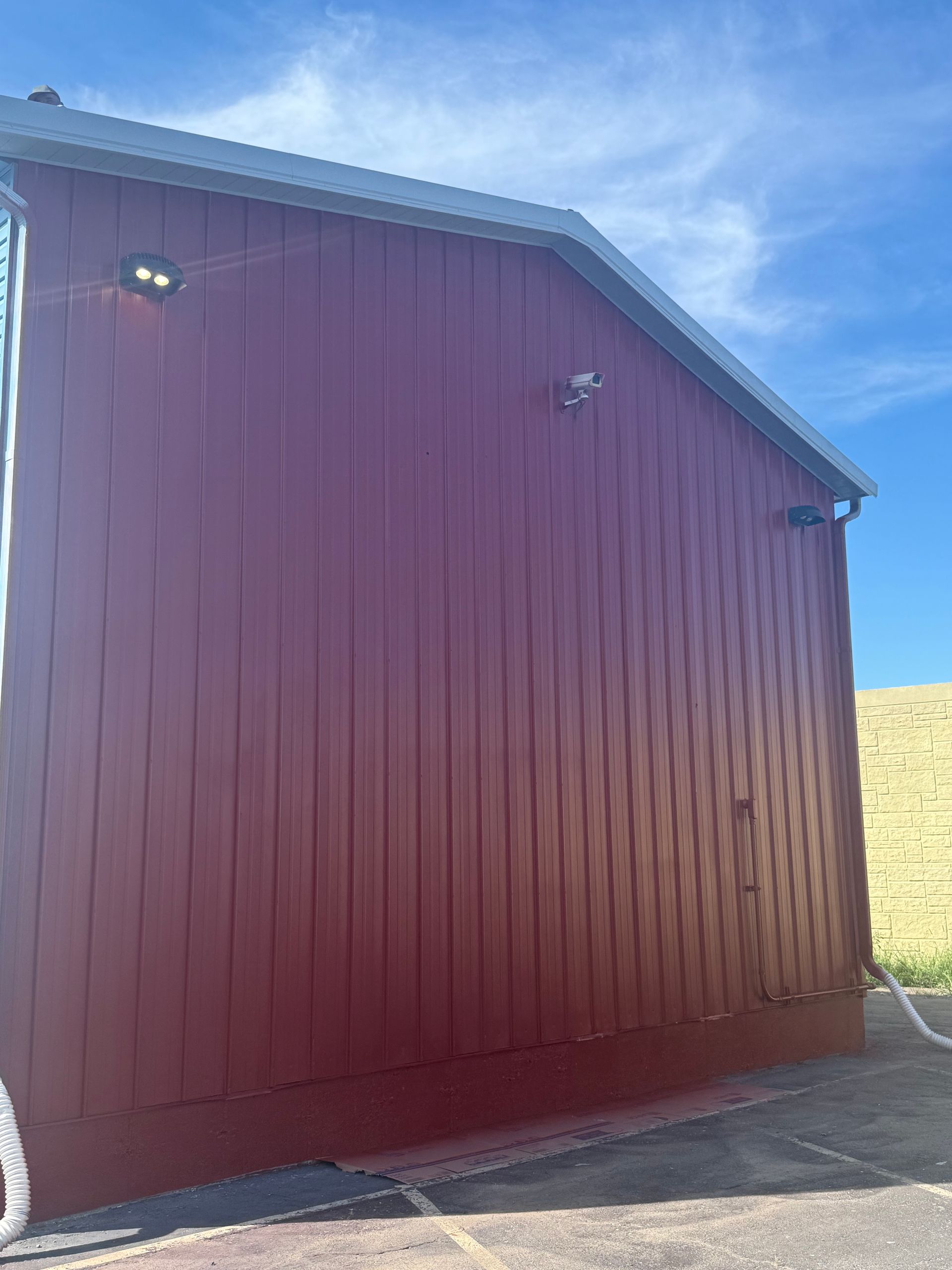 A low-angle view of a red, vertical-sided building against a blue sky, featuring exterior lights and a corner downspout.