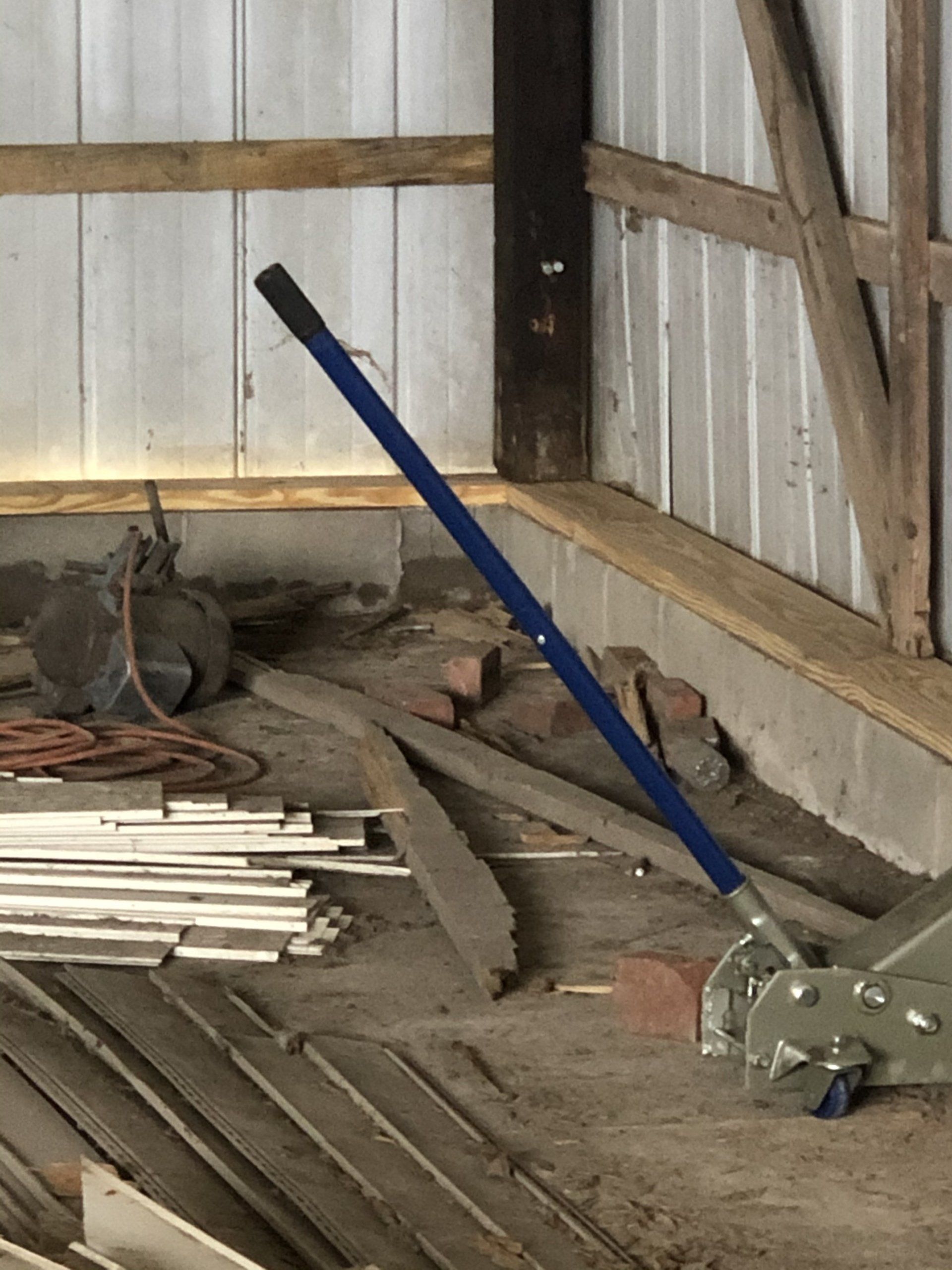 A blue-handled floor jack sits on a dirt floor in a shed near stacks of lumber.