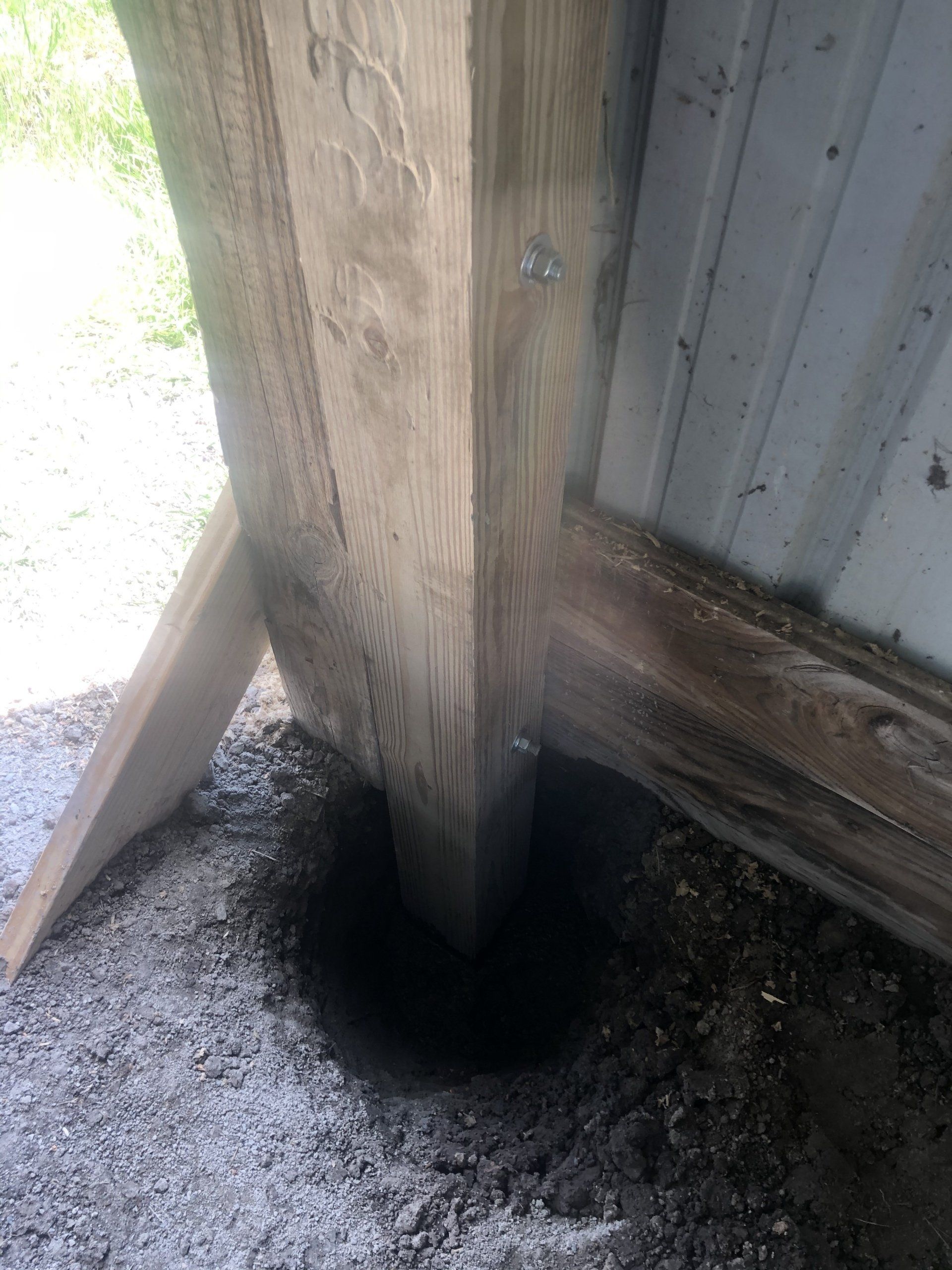 A wooden post stands in a newly dug hole in the ground next to a corrugated metal wall.