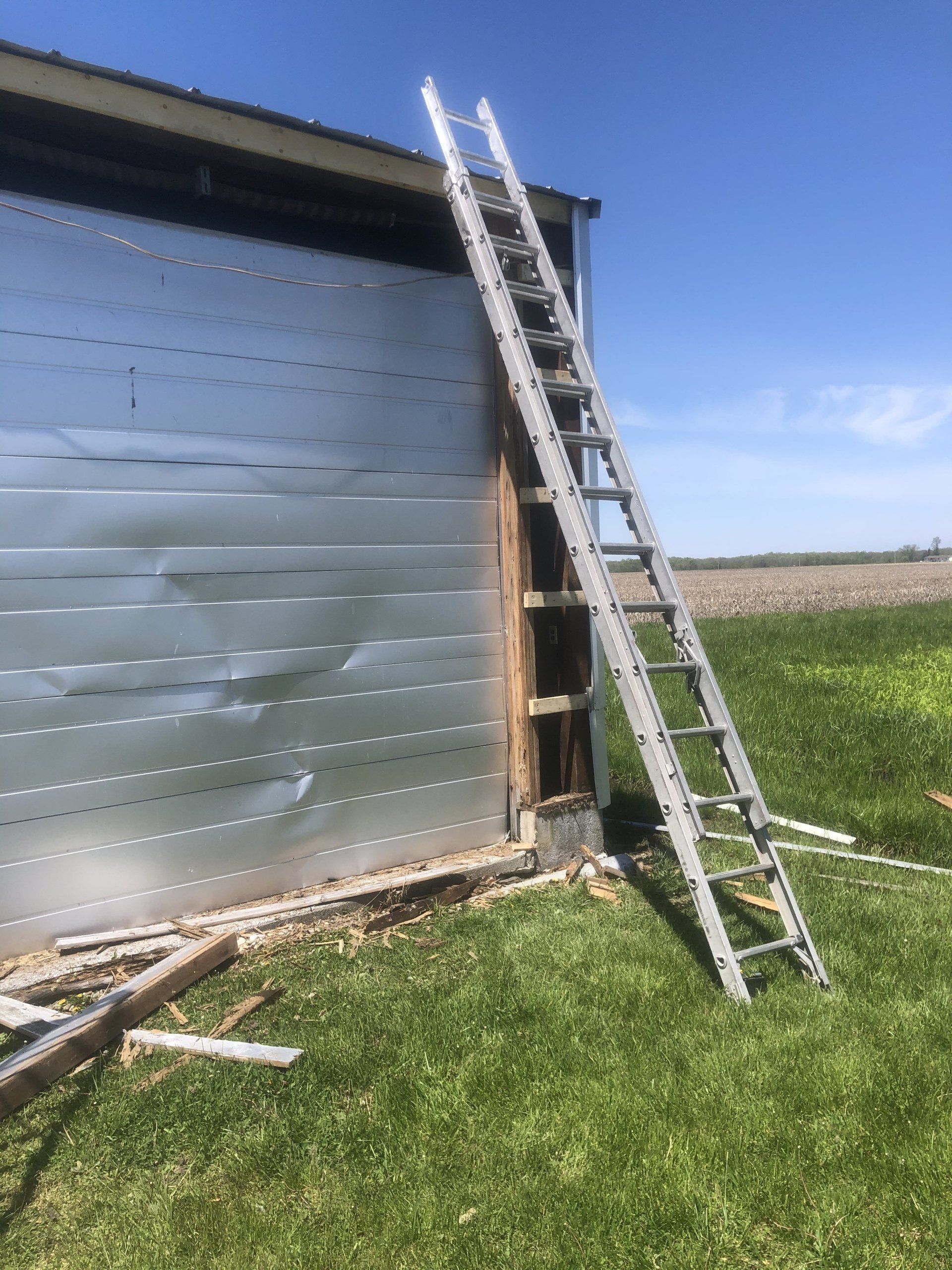 An extension ladder leans against the corner of a damaged metal-sided structure in a grassy field under a blue sky.