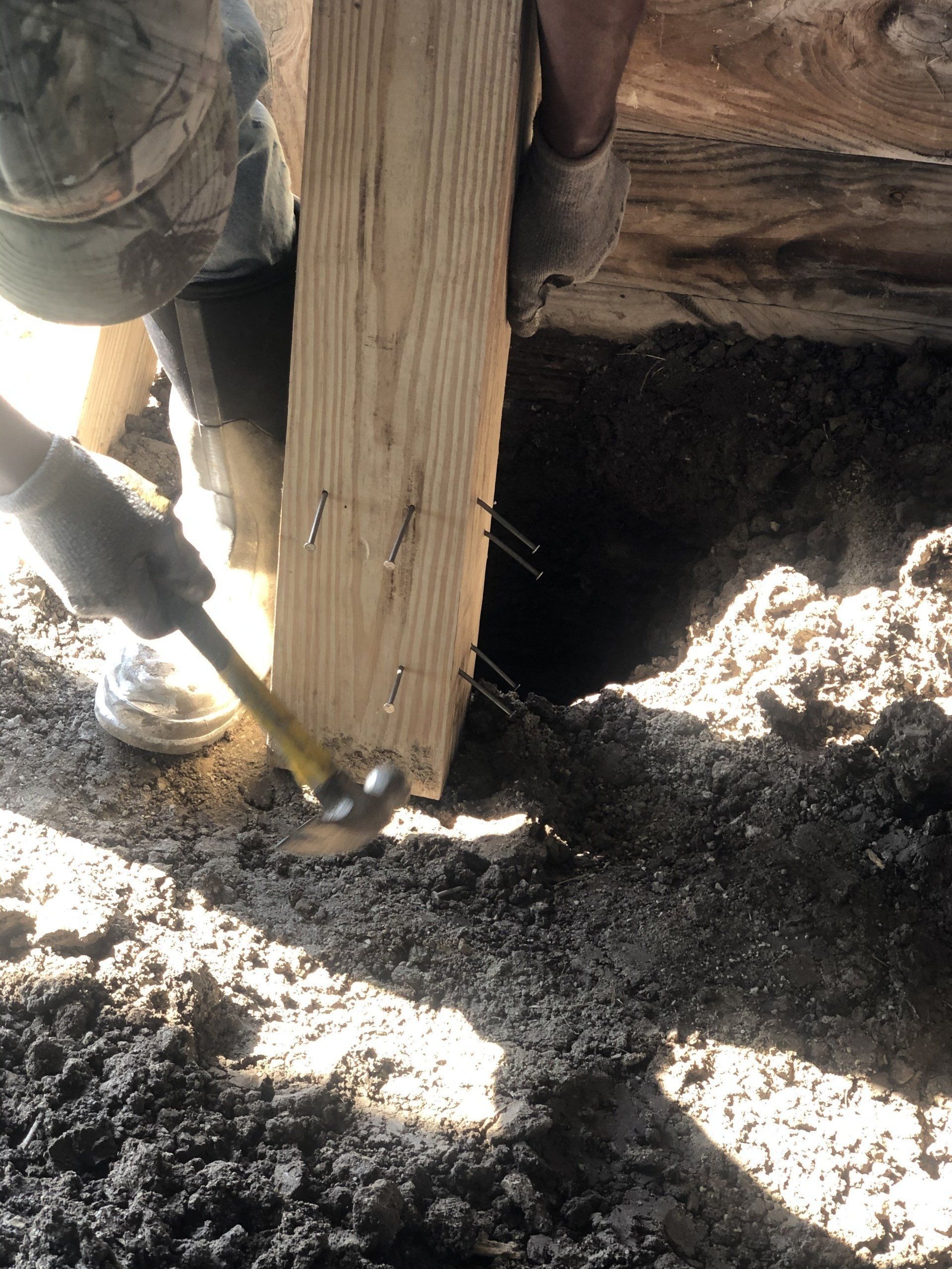 A person in work gloves uses a hammer to drive metal pins into a vertical wooden post set in a dark, excavated dirt hole.