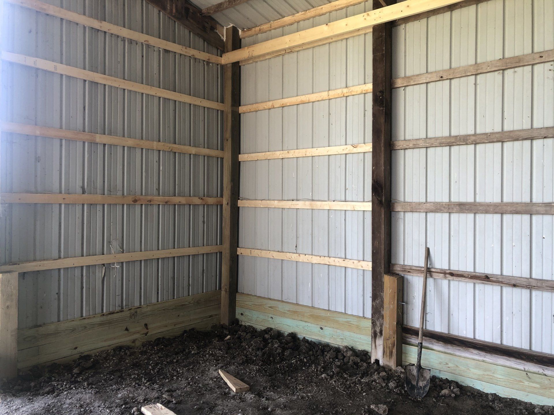 Interior corner of a metal-walled building showing vertical wooden posts, horizontal girts, and a dirt floor.