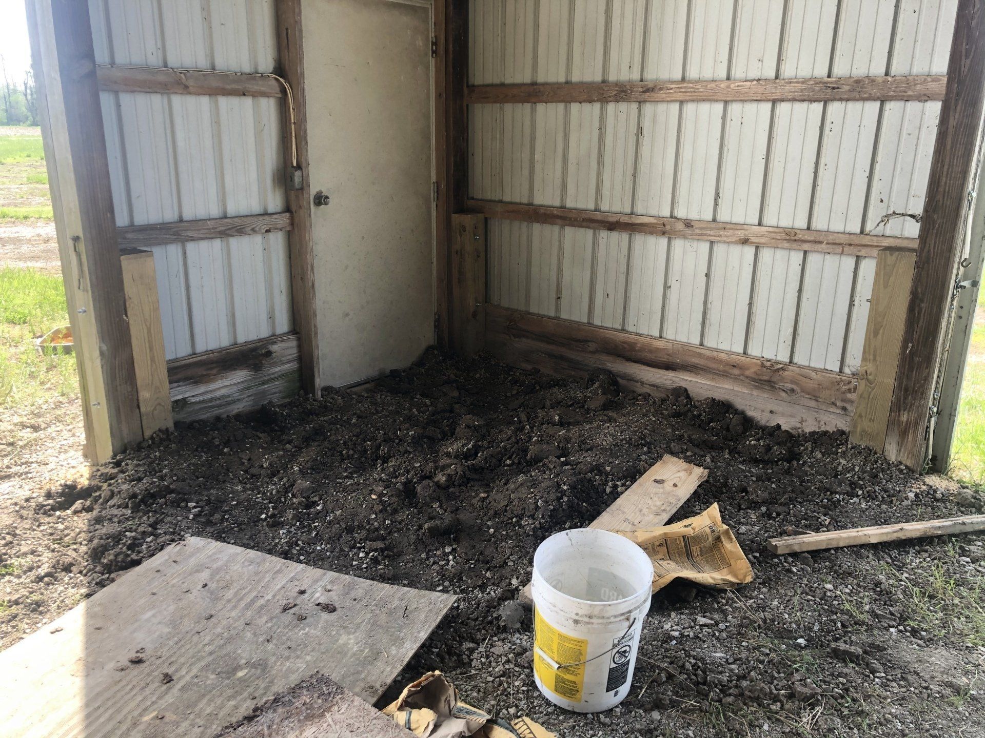 A dirt floor inside a metal shed with a wooden door and a white plastic bucket sitting on the ground.