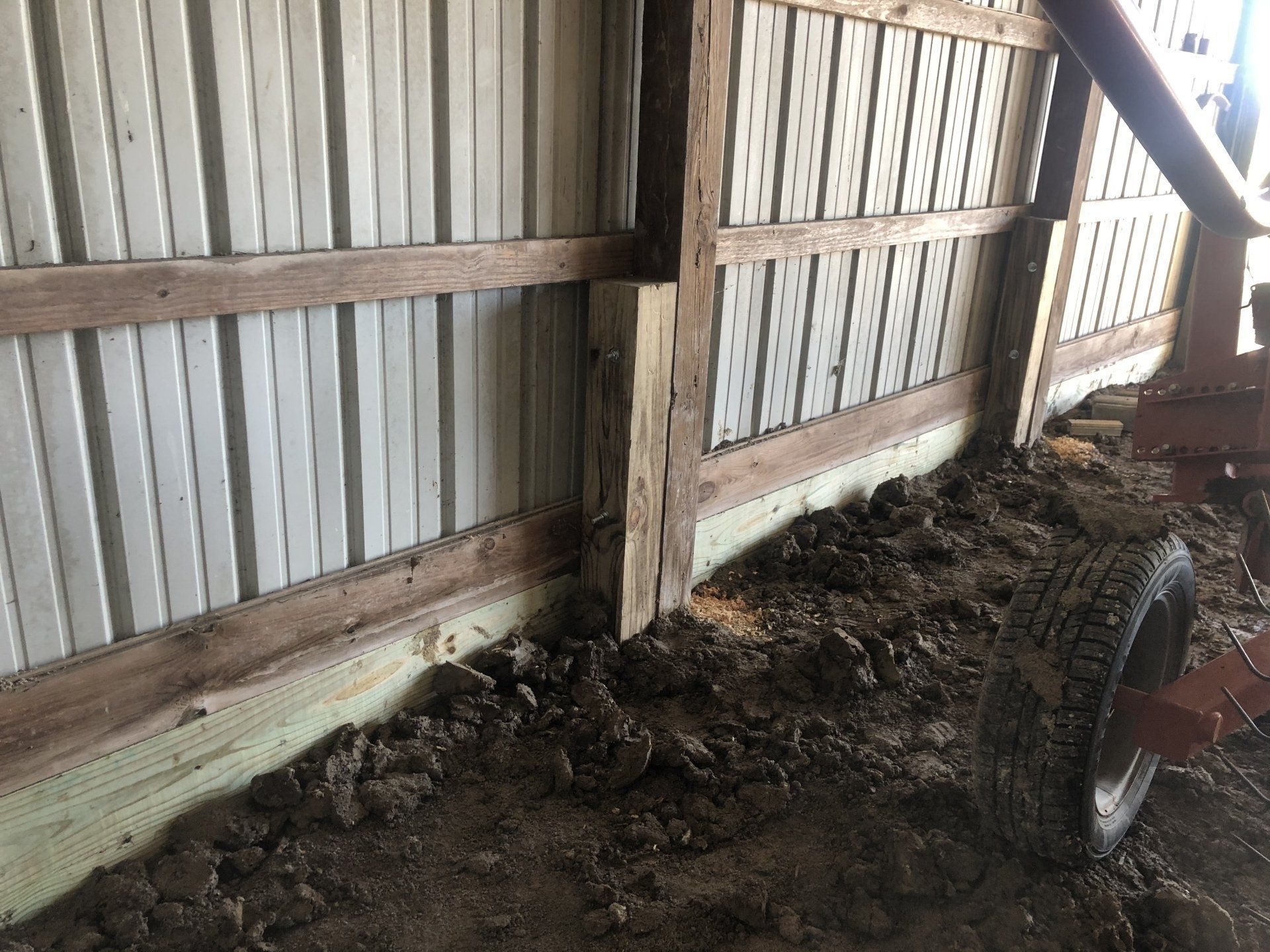 The interior wall of a pole barn with corrugated metal siding, wooden support posts, and an exposed dirt floor.