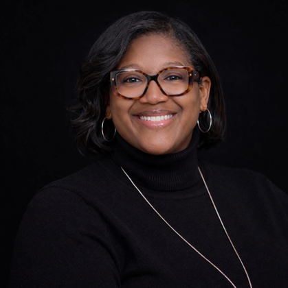 Smiling person with glasses and hoop earrings wearing a black turtleneck on a dark background