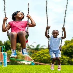 Two Black children happily swinging on a playground swing set in a grassy park. One girl laughs, the boy smiles.